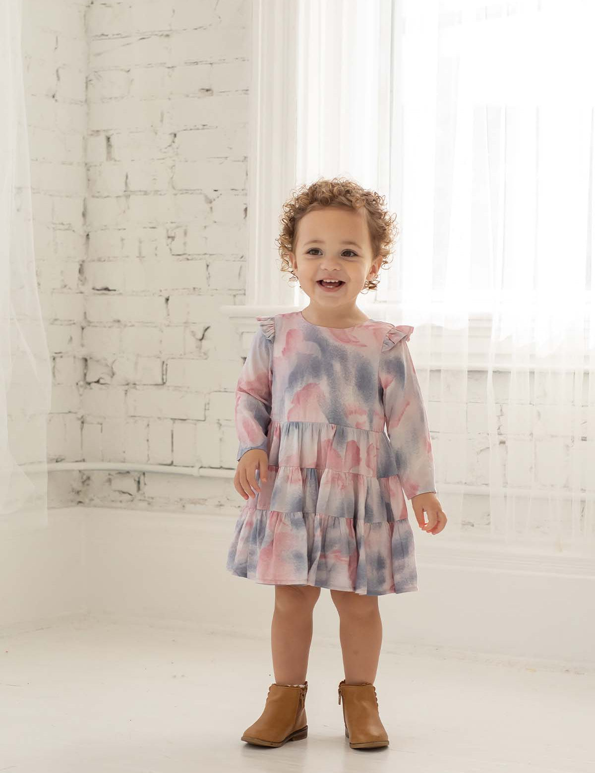 A smiling toddler with curly hair wears the Mabel + Honey Purple Haze Rayon Dress, featuring long sleeves and a tiered tie-dye design, standing in a bright white room with sheer curtains and a painted brick wall.