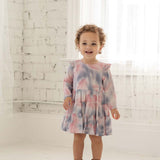 A smiling toddler with curly hair wears the Mabel + Honey Purple Haze Rayon Dress, featuring long sleeves and a tiered tie-dye design, standing in a bright white room with sheer curtains and a painted brick wall.