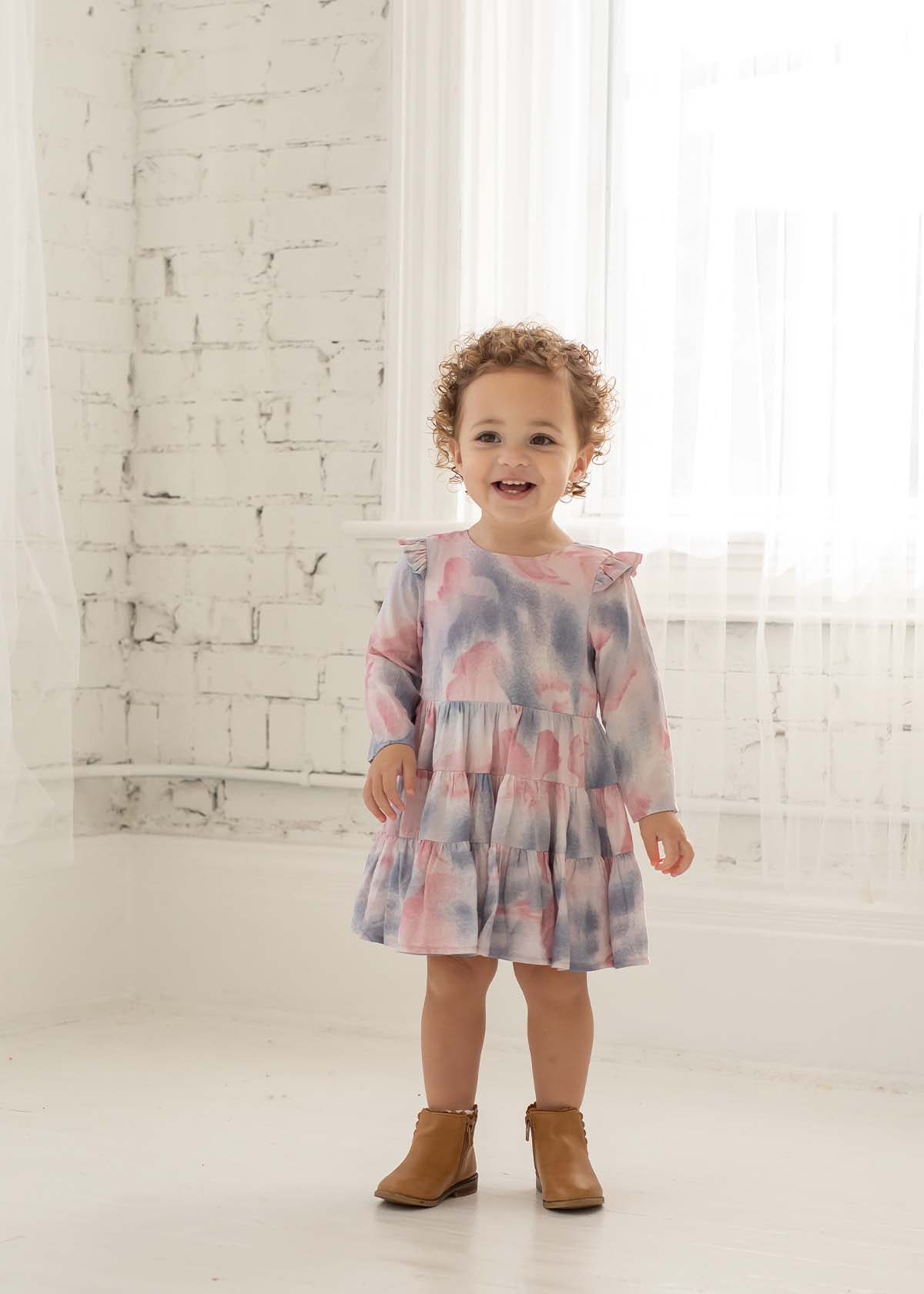 A smiling toddler with curly hair wears the Mabel + Honey Purple Haze Rayon Dress, featuring long sleeves and a tiered tie-dye design, standing in a bright white room with sheer curtains and a painted brick wall.