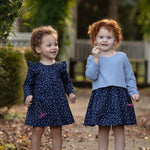 Two young children with curly hair stand outdoors on a path, each wearing the Midnight Woven Dress by Mabel + Honey in matching blue polka dots with charming ruffle cap sleeves. Smiling and holding small flowers, they are surrounded by green foliage and fallen leaves.