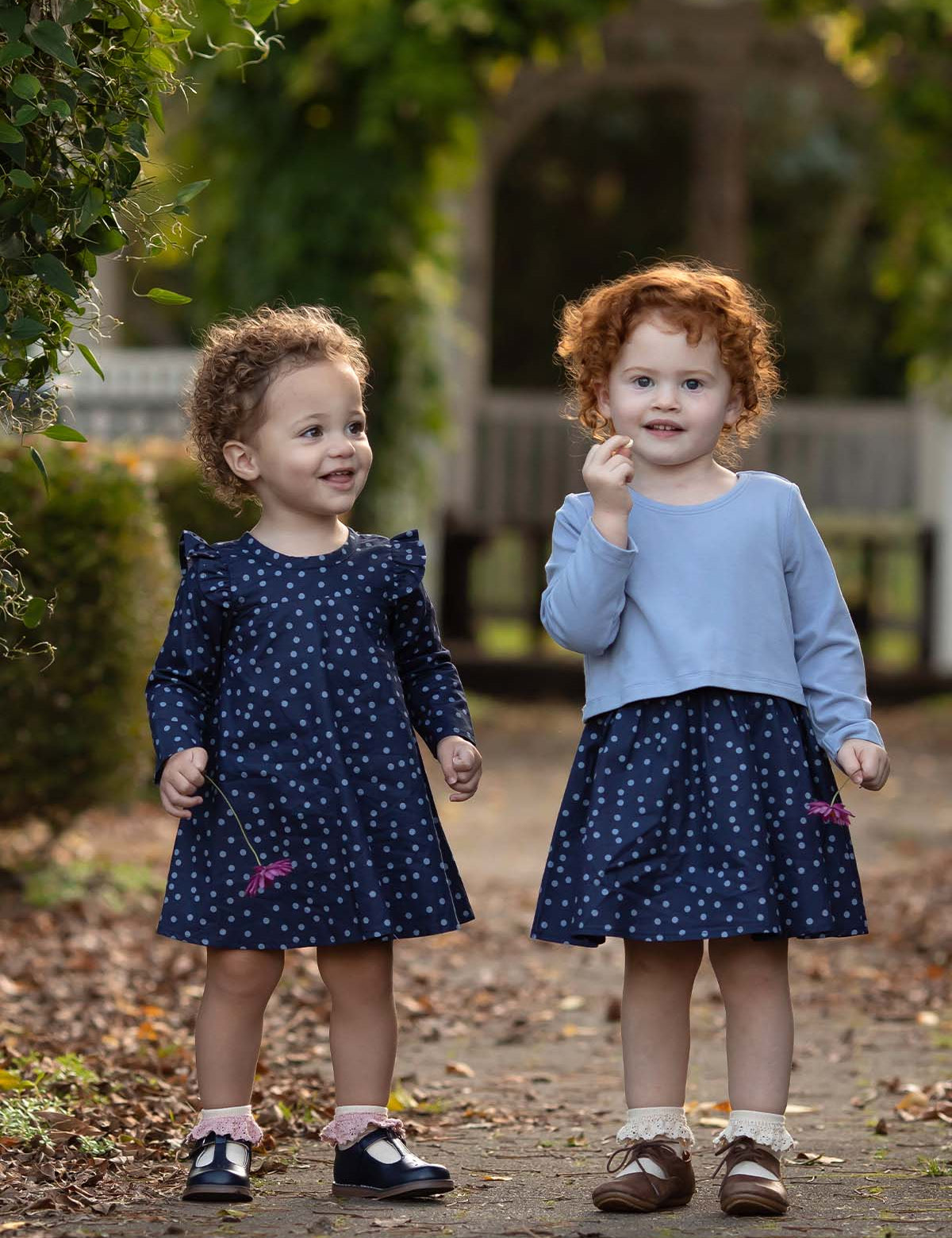 Two young children with curly hair stand outdoors on a path, each wearing the Midnight Woven Dress by Mabel + Honey in matching blue polka dots with charming ruffle cap sleeves. Smiling and holding small flowers, they are surrounded by green foliage and fallen leaves.