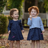 Two young children with curly hair stand outdoors on a path, each wearing the Midnight Woven Dress by Mabel + Honey in matching blue polka dots with charming ruffle cap sleeves. Smiling and holding small flowers, they are surrounded by green foliage and fallen leaves.