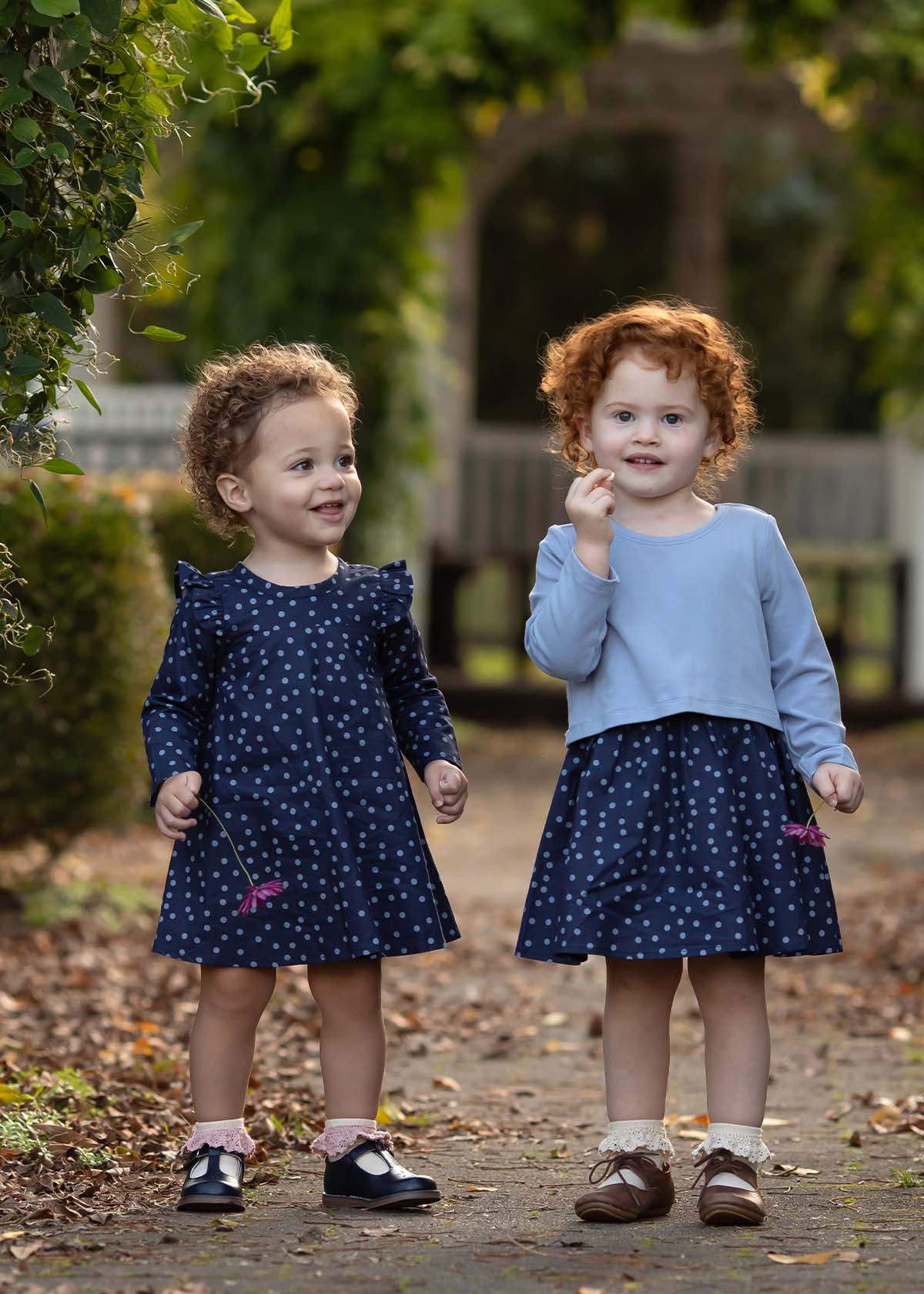 Two young children with curly hair stand outdoors on a path, each wearing the Midnight Woven Dress by Mabel + Honey in matching blue polka dots with charming ruffle cap sleeves. Smiling and holding small flowers, they are surrounded by green foliage and fallen leaves.
