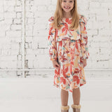 A smiling young girl with long blonde hair stands in front of a white brick wall, wearing the Mabel + Honey Water Color Rayon Dress—a long sleeve floral dress—paired with tan fur-trimmed boots.