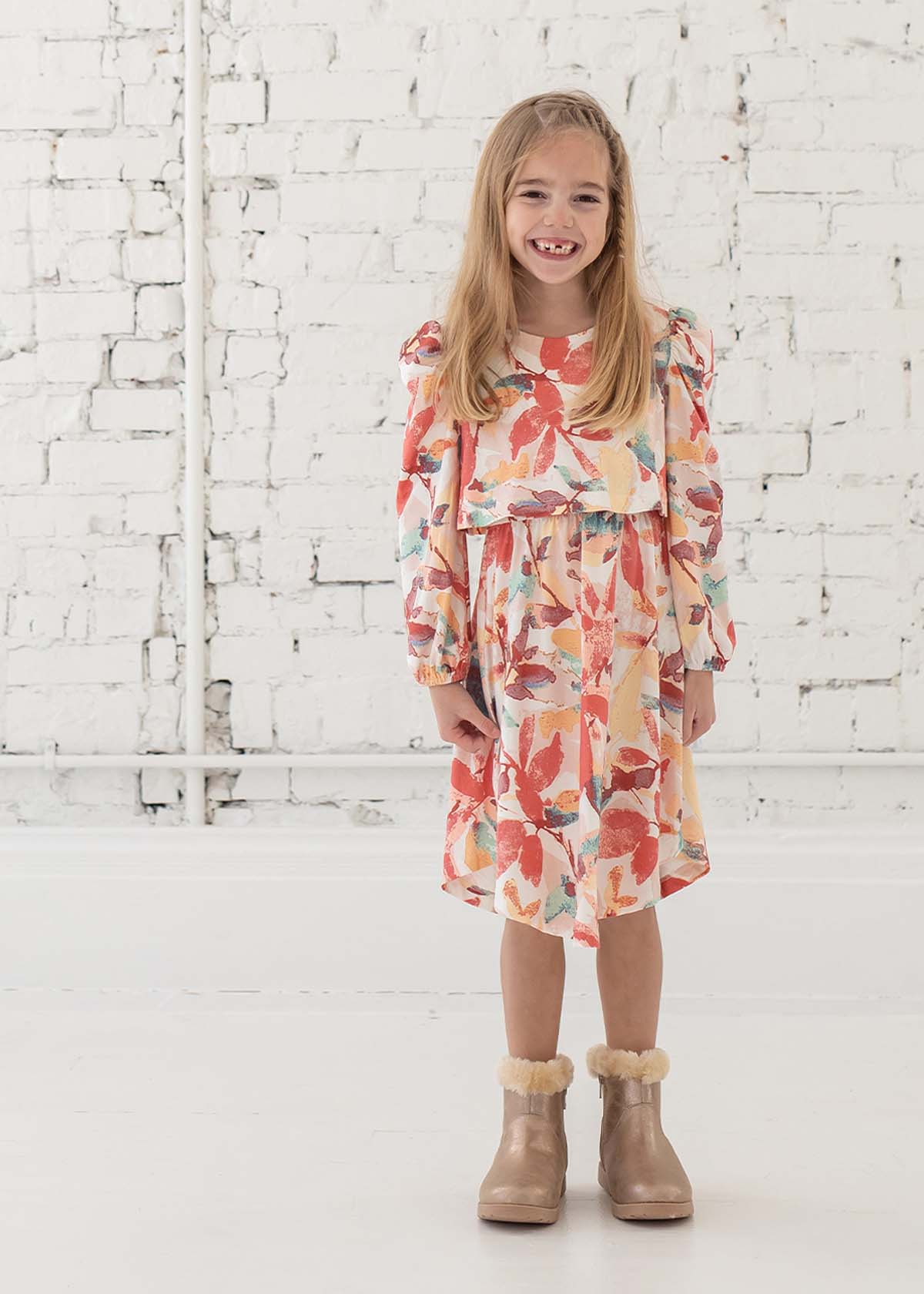 A smiling young girl with long blonde hair stands in front of a white brick wall, wearing the Mabel + Honey Water Color Rayon Dress—a long sleeve floral dress—paired with tan fur-trimmed boots.