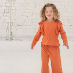 A young child with curly hair smiles indoors, dressed in the Mabel + Honey Candy Land Knit Two Piece Set featuring a pumpkin orange long-sleeve ruffle top and matching jogger pants, paired with white sneakers. The scene is set against a white brick wall and floor.