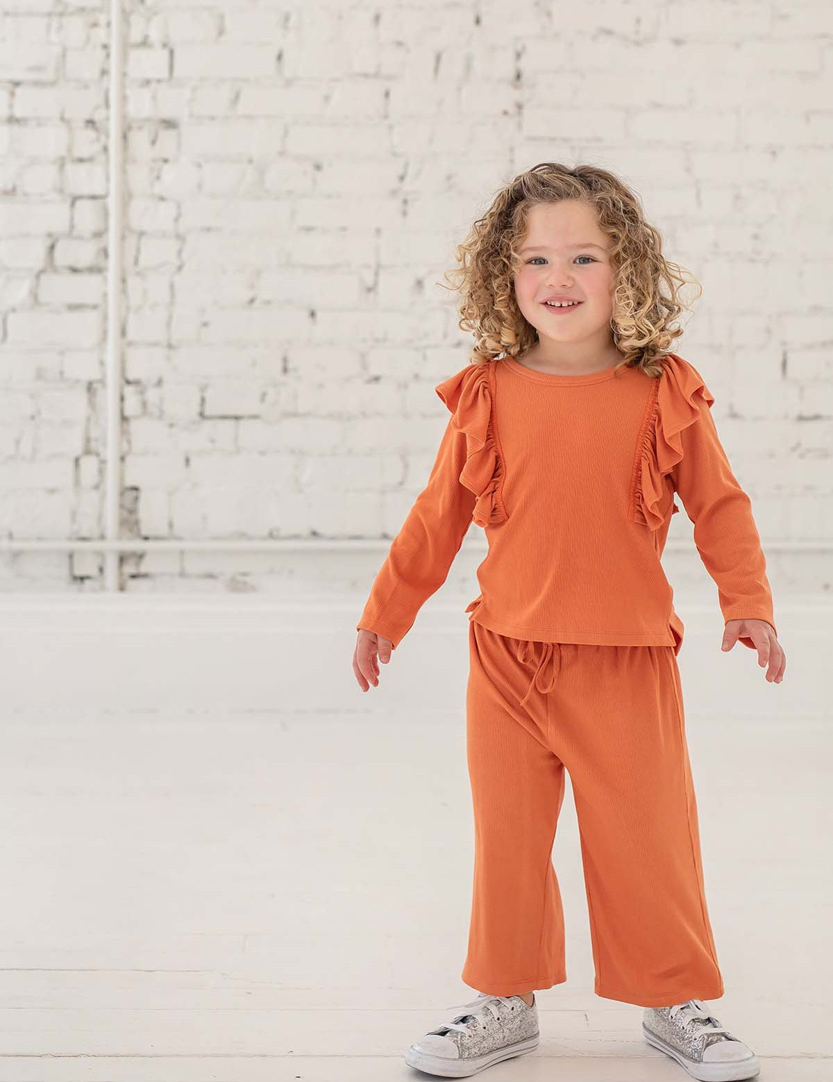 A young child with curly hair smiles indoors, dressed in the Mabel + Honey Candy Land Knit Two Piece Set featuring a pumpkin orange long-sleeve ruffle top and matching jogger pants, paired with white sneakers. The scene is set against a white brick wall and floor.