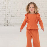 A young child with curly hair smiles indoors, dressed in the Mabel + Honey Candy Land Knit Two Piece Set featuring a pumpkin orange long-sleeve ruffle top and matching jogger pants, paired with white sneakers. The scene is set against a white brick wall and floor.