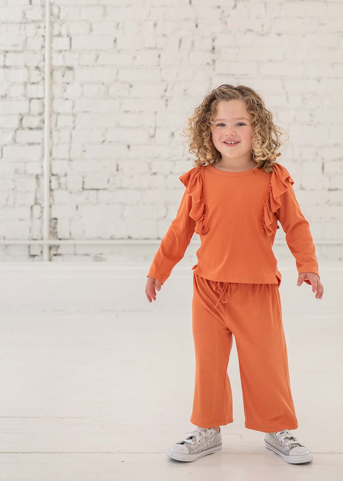 A young child with curly hair smiles indoors, dressed in the Mabel + Honey Candy Land Knit Two Piece Set featuring a pumpkin orange long-sleeve ruffle top and matching jogger pants, paired with white sneakers. The scene is set against a white brick wall and floor.
