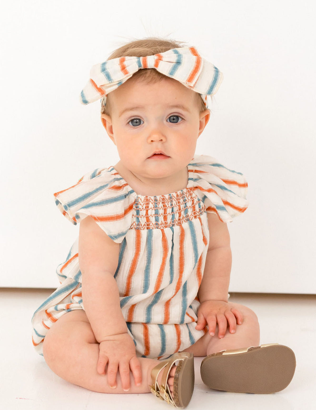 A baby wearing a Mabel and Honey Hermosa Headband and a soft cotton striped romper sits on the floor against a white background, gazing at the camera with a neutral expression.