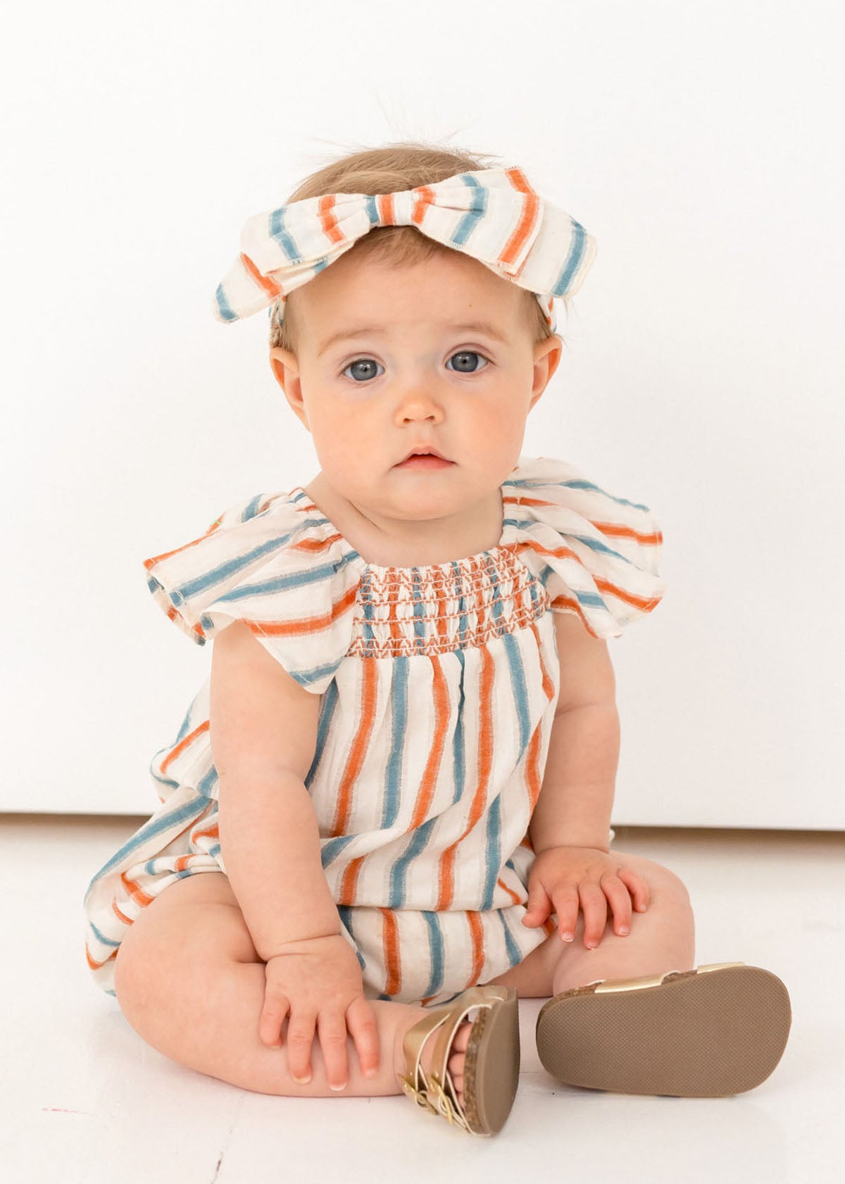 A baby wearing a Mabel and Honey Hermosa Headband and a soft cotton striped romper sits on the floor against a white background, gazing at the camera with a neutral expression.
