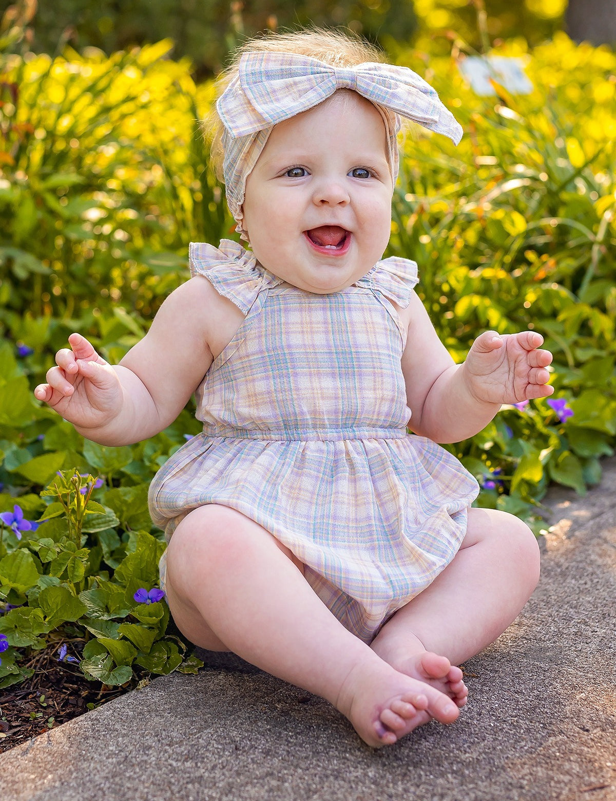 A smiling baby wears a Mabel and Honey Flower Farm Gingham Headband and matching romper, sitting barefoot on a stone path among green plants and small purple flowers in a sunlit garden.