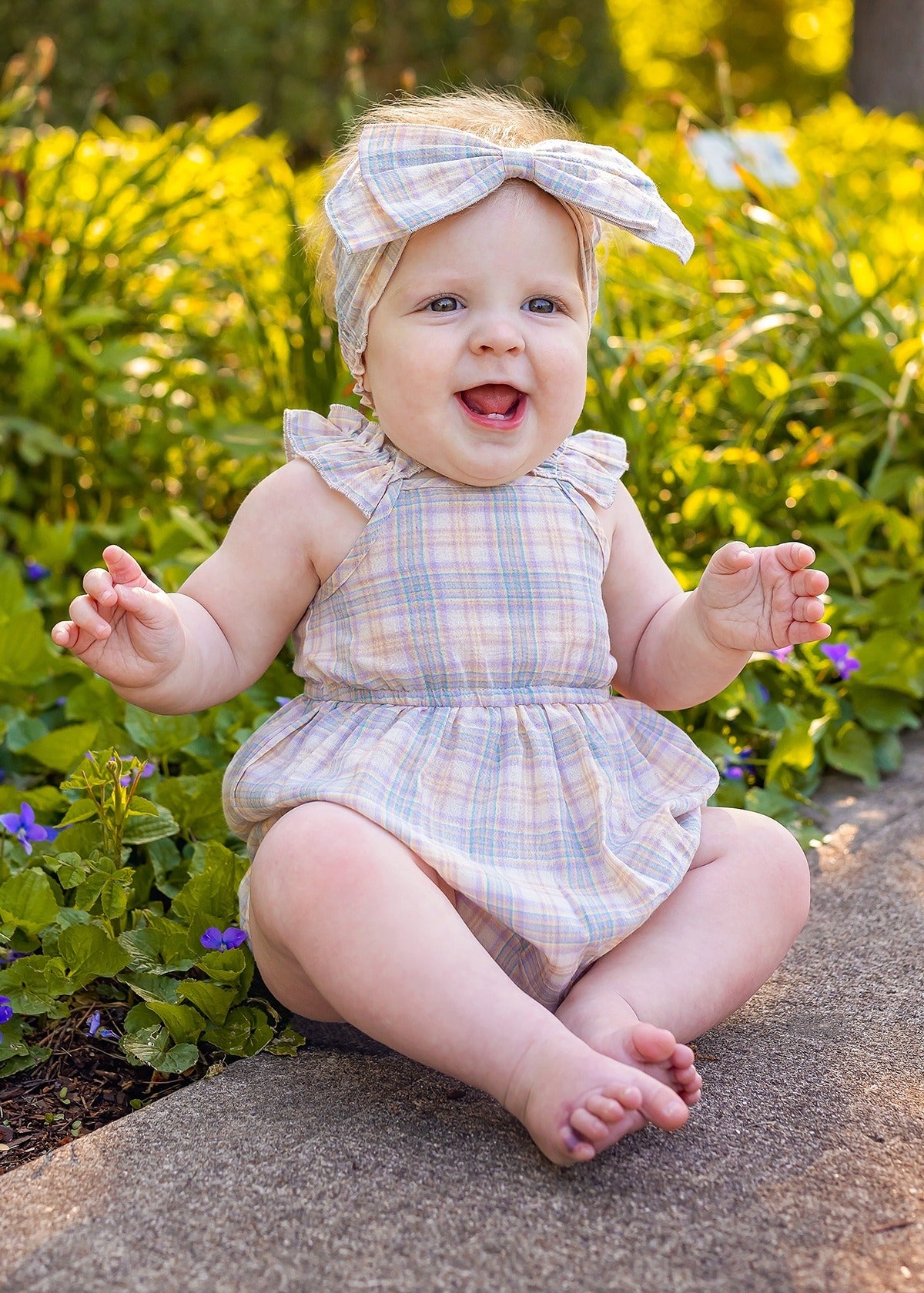 A smiling baby wears a Mabel and Honey Flower Farm Gingham Headband and matching romper, sitting barefoot on a stone path among green plants and small purple flowers in a sunlit garden.