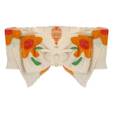 The Pleated Petals Headband by Mabel and Honey features cream-colored, pleated 100% cotton with vibrant orange and yellow floral prints, green leaves, and a bold front bow for a striking decorative look.