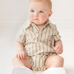 A baby with light hair and blue eyes sits on the floor in a Mabel and Honey Meadow Breeze Striped Baby Romper with white shoes, gazing to the side in a bright, softly lit room.