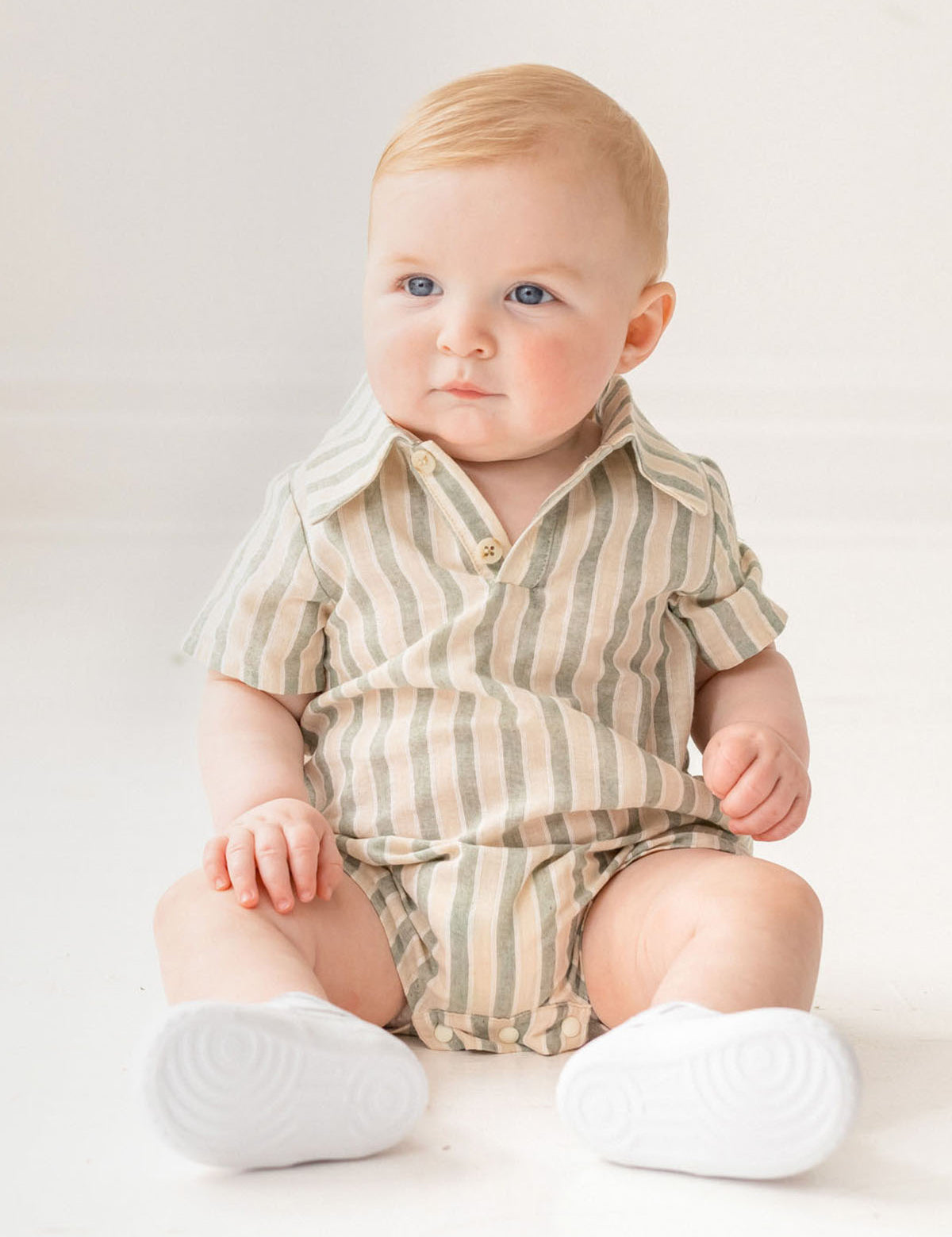 A baby with light hair and blue eyes sits on the floor in a Mabel and Honey Meadow Breeze Striped Baby Romper with white shoes, gazing to the side in a bright, softly lit room.
