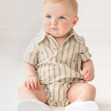 A baby with light hair and blue eyes sits on the floor in a Mabel and Honey Meadow Breeze Striped Baby Romper with white shoes, gazing to the side in a bright, softly lit room.