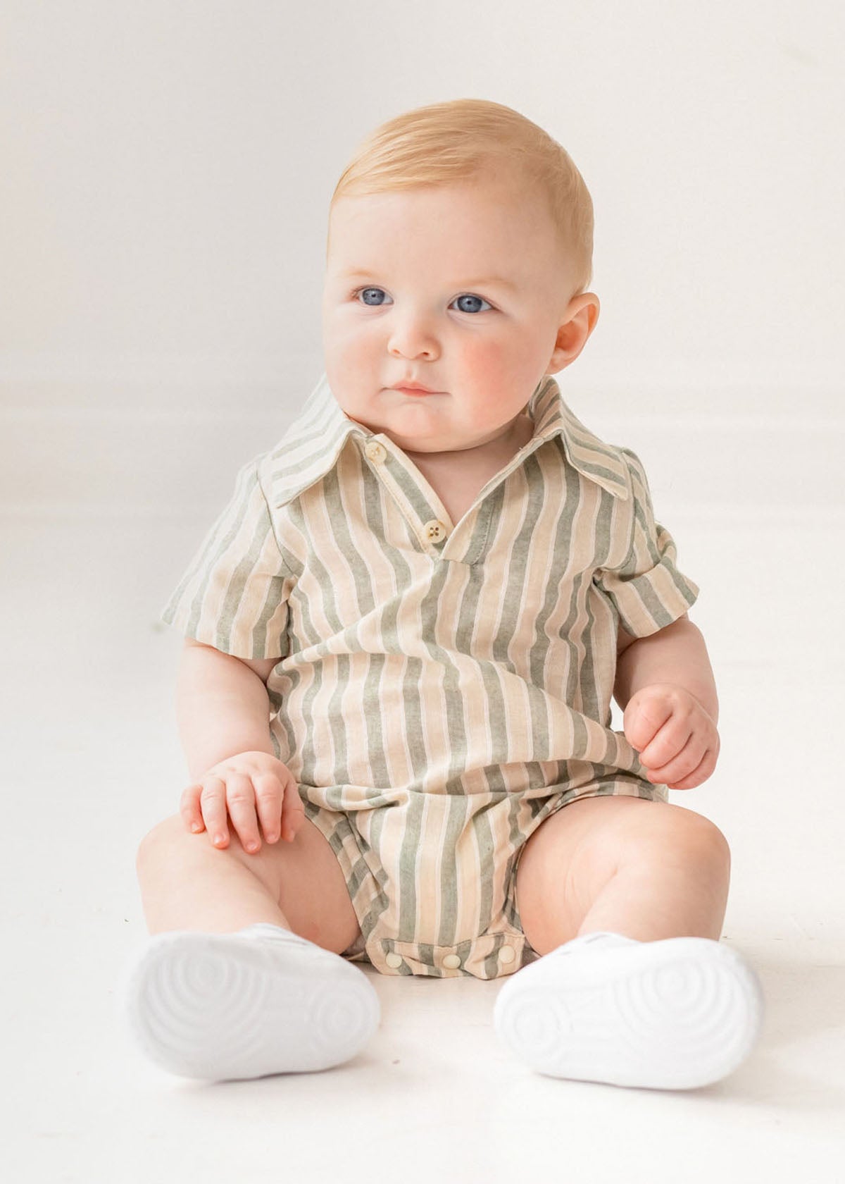 A baby with light hair and blue eyes sits on the floor in a Mabel and Honey Meadow Breeze Striped Baby Romper with white shoes, gazing to the side in a bright, softly lit room.