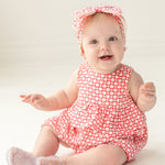 A smiling baby with light hair sits on a light-colored floor, wearing the Mabel and Honey Little Red Wagon Baby Romper with a matching headband and translucent socks, looking up.
