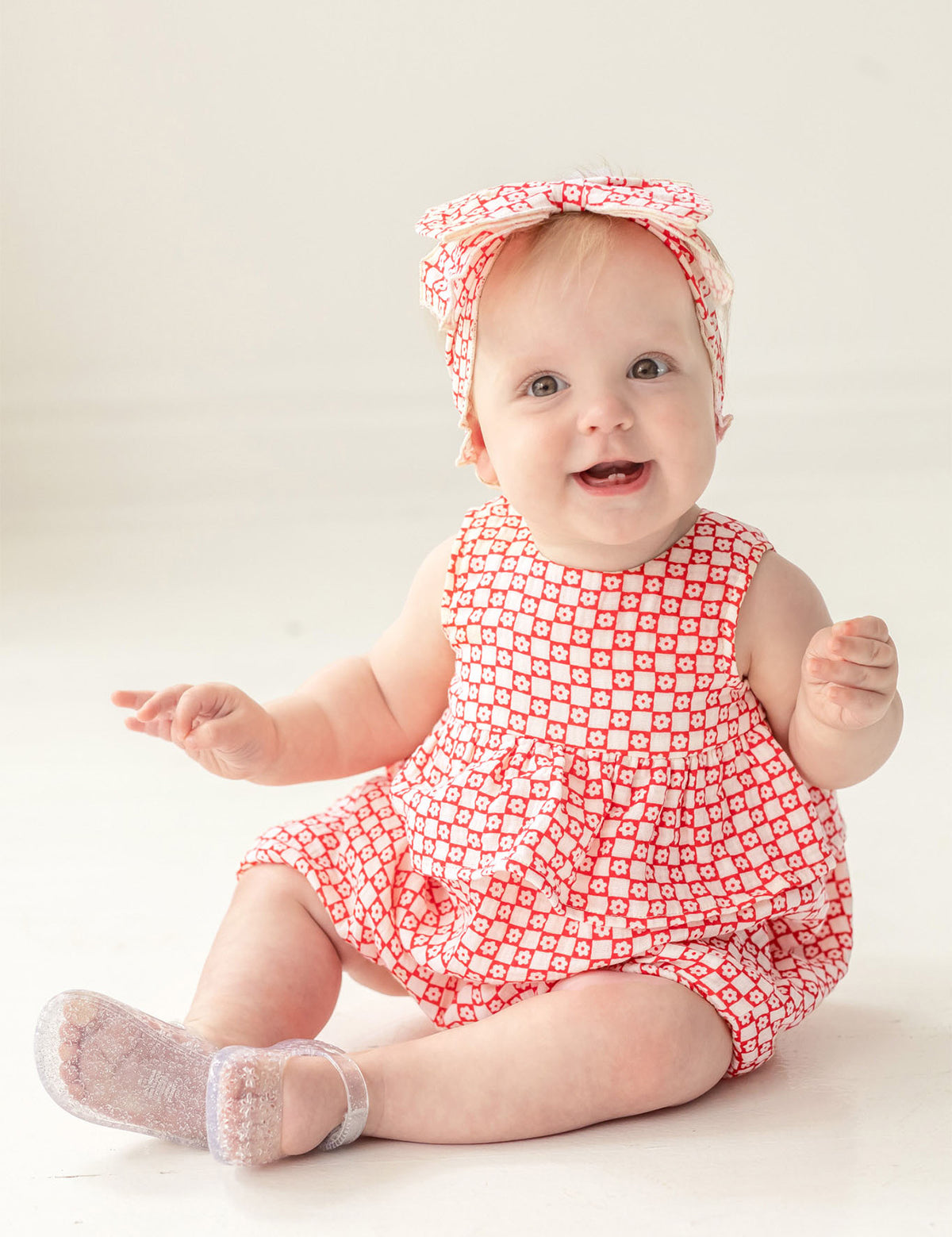 A smiling baby with light hair sits on a light-colored floor, wearing the Mabel and Honey Little Red Wagon Baby Romper with a matching headband and translucent socks, looking up.