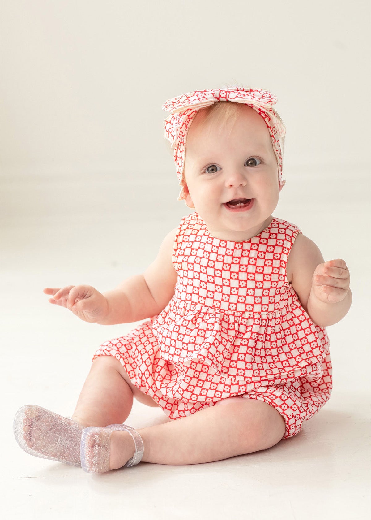 A smiling baby with light hair sits on a light-colored floor, wearing the Mabel and Honey Little Red Wagon Baby Romper with a matching headband and translucent socks, looking up.