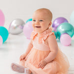 A happy baby wears the Isobella and Chloe Creamsicle Sleeveless Dress, sitting on a white floor amid purple, teal, and silver balloons. Soft natural light highlights her smile as she enjoys her special moment.
