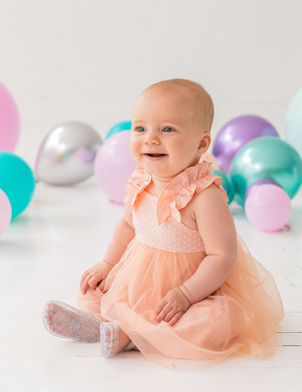 A happy baby wears the Isobella and Chloe Creamsicle Sleeveless Dress, sitting on a white floor amid purple, teal, and silver balloons. Soft natural light highlights her smile as she enjoys her special moment.