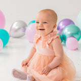 A happy baby wears the Isobella and Chloe Creamsicle Sleeveless Dress, sitting on a white floor amid purple, teal, and silver balloons. Soft natural light highlights her smile as she enjoys her special moment.