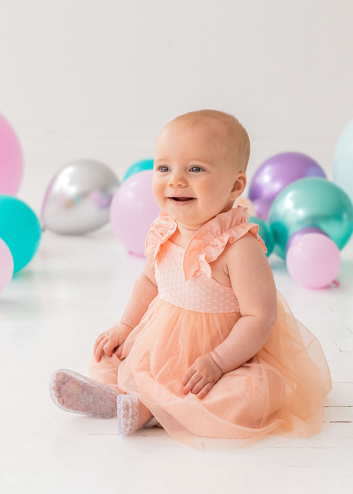 A happy baby wears the Isobella and Chloe Creamsicle Sleeveless Dress, sitting on a white floor amid purple, teal, and silver balloons. Soft natural light highlights her smile as she enjoys her special moment.