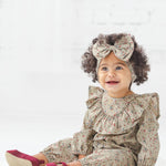A smiling toddler with curly hair sits on a white floor in a bright room, wearing the Mabel + Honey Berry & Olive Wreath Rayon Romper with its charming ruffle collar, paired with a matching headband featuring a large bow and maroon shoes.