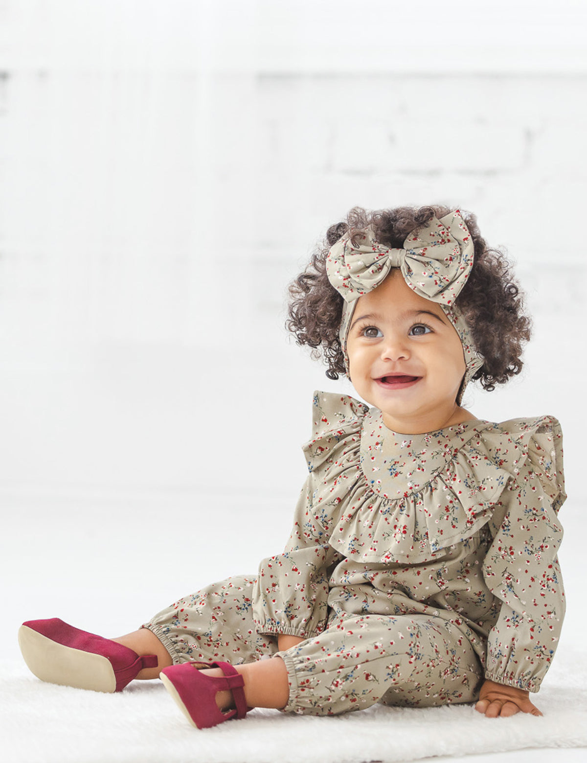 A smiling toddler with curly hair sits on a white floor in a bright room, wearing the Mabel + Honey Berry & Olive Wreath Rayon Romper with its charming ruffle collar, paired with a matching headband featuring a large bow and maroon shoes.