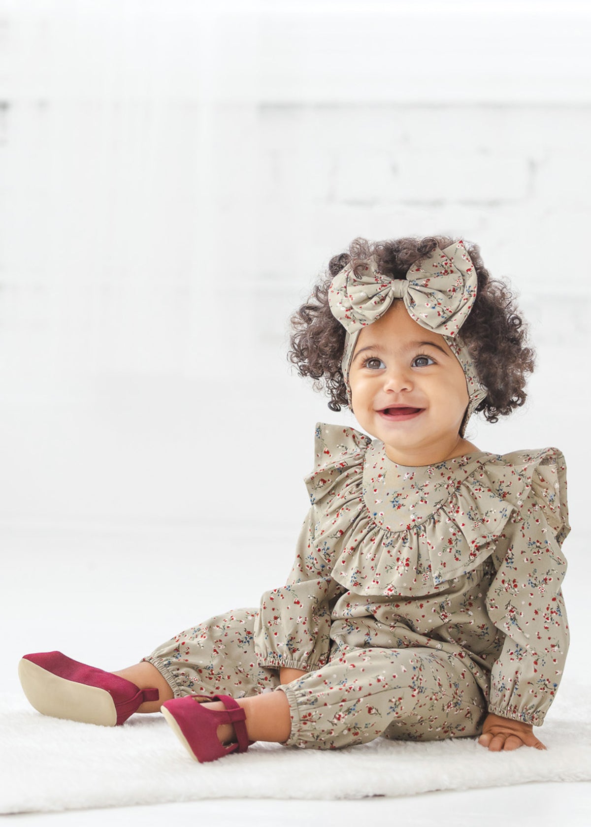 A smiling toddler with curly hair sits on a white floor in a bright room, wearing the Mabel + Honey Berry & Olive Wreath Rayon Romper with its charming ruffle collar, paired with a matching headband featuring a large bow and maroon shoes.