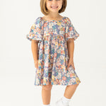 A young girl with short light brown hair smiles against a white background, wearing the Mabel and Honey Flower Mae Floral Dress with short sleeves, paired with white socks and shoes.