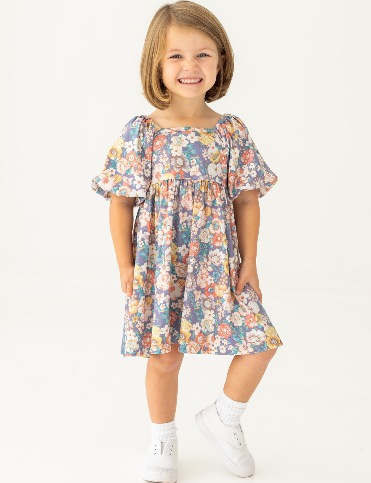A young girl with short light brown hair smiles against a white background, wearing the Mabel and Honey Flower Mae Floral Dress with short sleeves, paired with white socks and shoes.