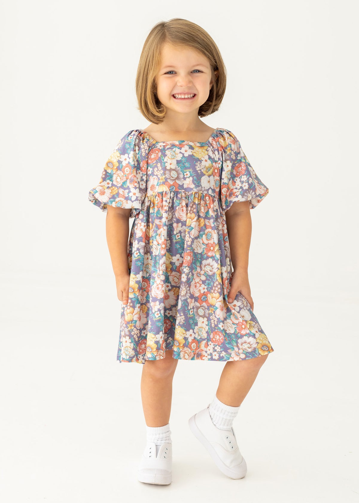 A young girl with short light brown hair smiles against a white background, wearing the Mabel and Honey Flower Mae Floral Dress with short sleeves, paired with white socks and shoes.