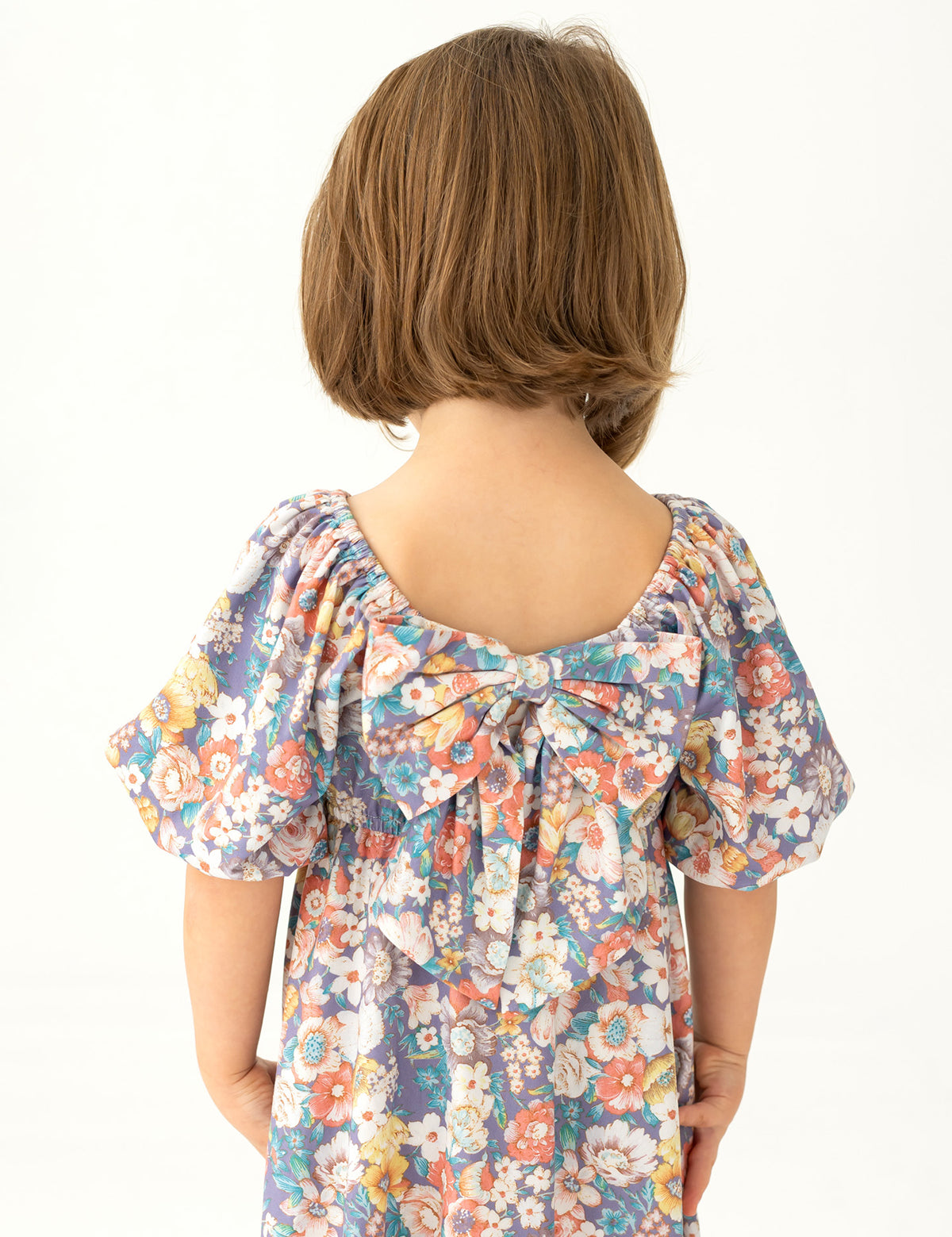 A young child with short brown hair is seen from behind, wearing the Mabel and Honey Flower Mae Floral Dress, featuring puffed sleeves, a floral print, and a large back bow. The background is plain and light-colored.