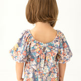A young child with short brown hair is seen from behind, wearing the Mabel and Honey Flower Mae Floral Dress, featuring puffed sleeves, a floral print, and a large back bow. The background is plain and light-colored.