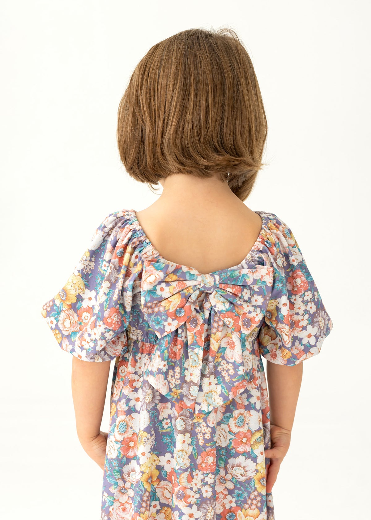 A young child with short brown hair is seen from behind, wearing the Mabel and Honey Flower Mae Floral Dress, featuring puffed sleeves, a floral print, and a large back bow. The background is plain and light-colored.