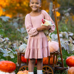 A smiling girl in the Mabel and Honey Flower Mae Pink Dress stands outdoors holding a chicken, surrounded by pumpkins and autumn foliage, with a wooden cart behind her for a cozy fall vibe.