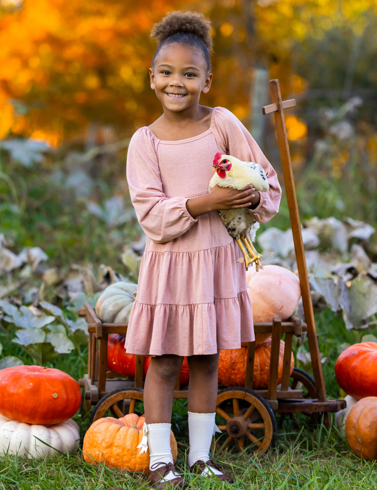 A smiling girl in the Mabel and Honey Flower Mae Pink Dress stands outdoors holding a chicken, surrounded by pumpkins and autumn foliage, with a wooden cart behind her for a cozy fall vibe.