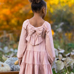 A young girl with curly hair in a bun stands outdoors among pumpkins and autumn leaves, wearing the Flower Mae Pink Dress by Mabel and Honey, featuring a large bow on the back.