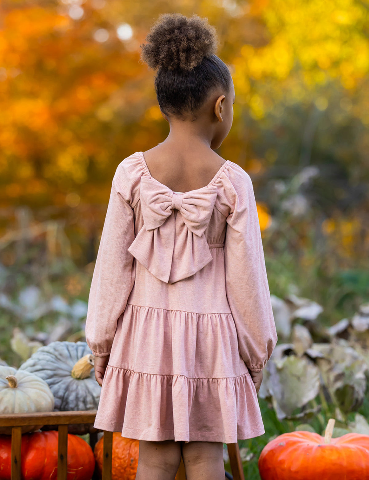 A young girl with curly hair in a bun stands outdoors among pumpkins and autumn leaves, wearing the Flower Mae Pink Dress by Mabel and Honey, featuring a large bow on the back.