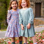 Two young girls stand on grass with red and white leaves. One wears the Mabel and Honey Organic Blooms Dress, the other a light blue dress. Both are smiling close together, with trees and a stone building in the background.