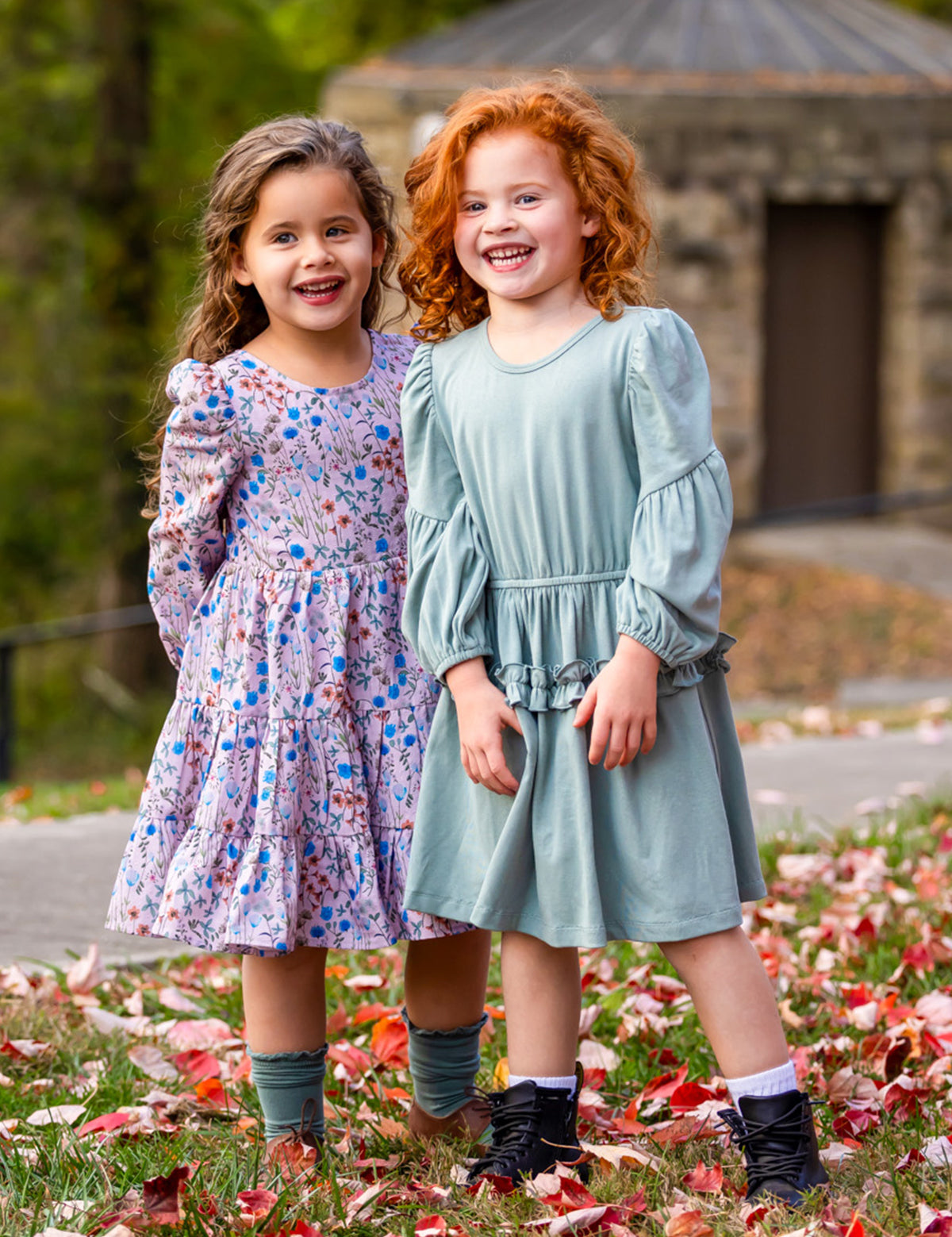 Two young girls stand on grass with red and white leaves. One wears the Mabel and Honey Organic Blooms Dress, the other a light blue dress. Both are smiling close together, with trees and a stone building in the background.