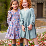 Two young girls stand on grass with red and white leaves. One wears the Mabel and Honey Organic Blooms Dress, the other a light blue dress. Both are smiling close together, with trees and a stone building in the background.