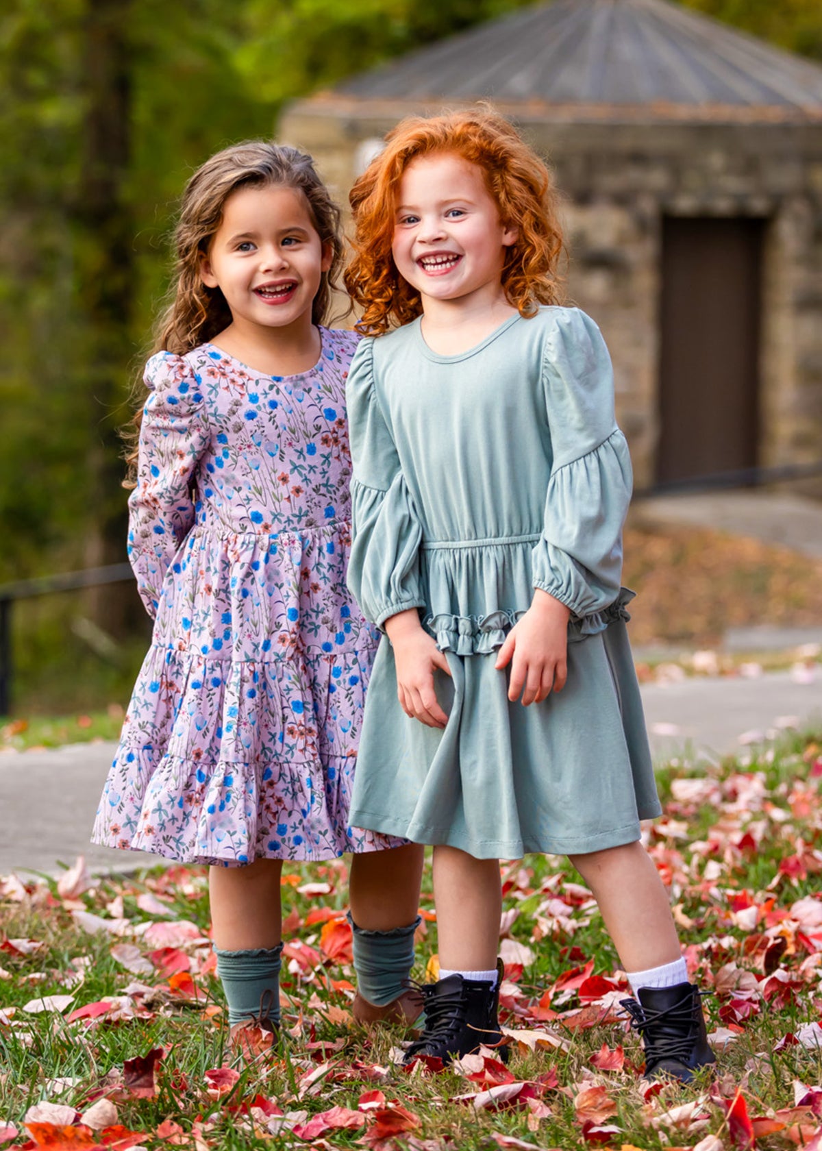 Two young girls stand on grass with red and white leaves. One wears the Mabel and Honey Organic Blooms Dress, the other a light blue dress. Both are smiling close together, with trees and a stone building in the background.