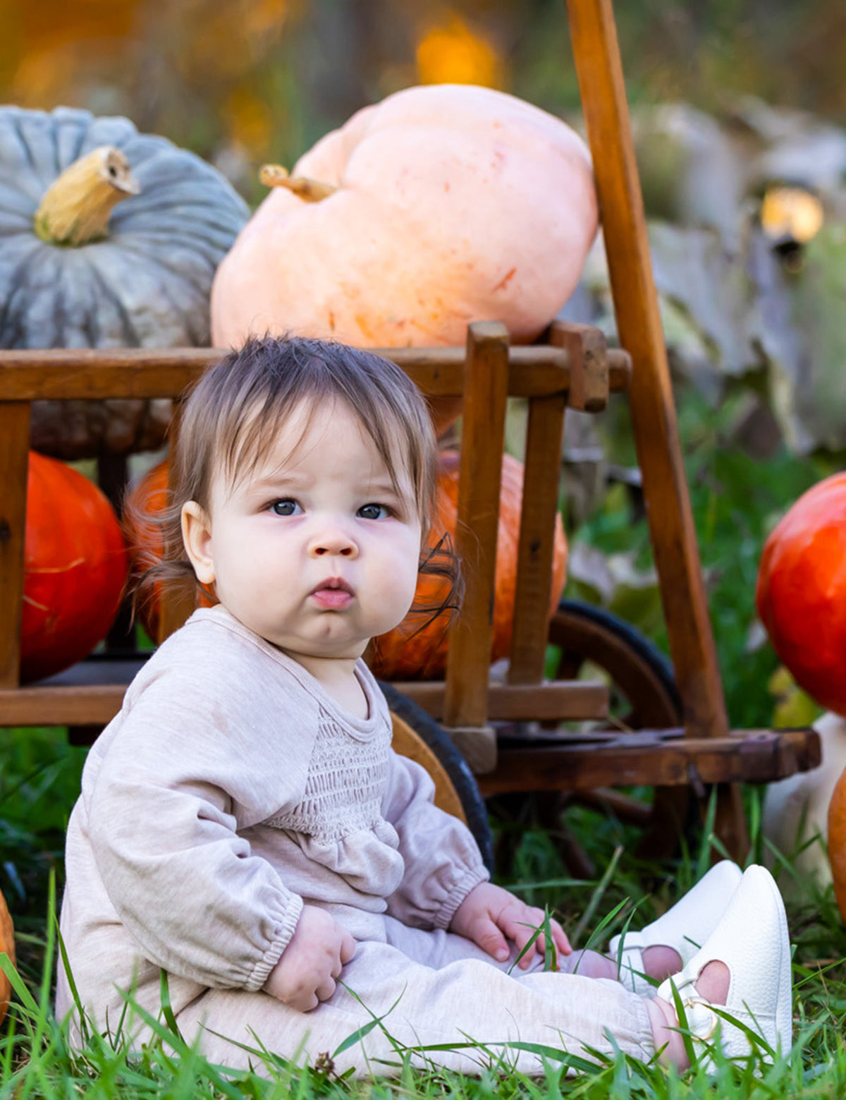 A baby wears the Millie Romper by Mabel and Honey while sitting on grass before a wooden cart of pumpkins and gourds, creating a cozy autumn scene with seasonal hues and greenery.