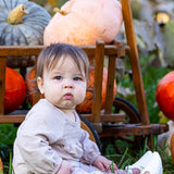 A baby wears the Millie Romper by Mabel and Honey while sitting on grass before a wooden cart of pumpkins and gourds, creating a cozy autumn scene with seasonal hues and greenery.