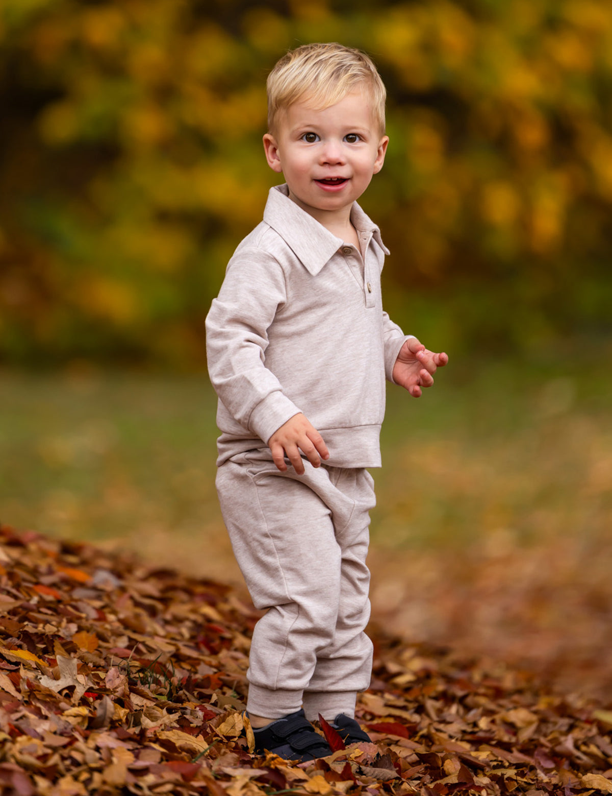 A young child with blonde hair stands on autumn leaves, wearing the Beckett and Bear Millie Brothers 2 PC Set with a collared neckline and dark shoes, set against a backdrop of blurred yellow and green foliage.