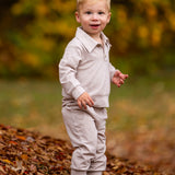 A young child with blonde hair stands on autumn leaves, wearing the Beckett and Bear Millie Brothers 2 PC Set with a collared neckline and dark shoes, set against a backdrop of blurred yellow and green foliage.